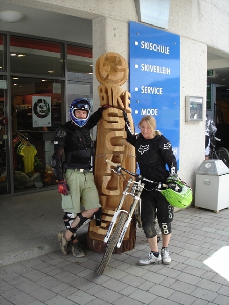 Bikeschul-Besitzerin Sabine Enzinger und ein weiterer Downhiller vor einer Holzschnitzerei mit der Inschrift Bikeschule